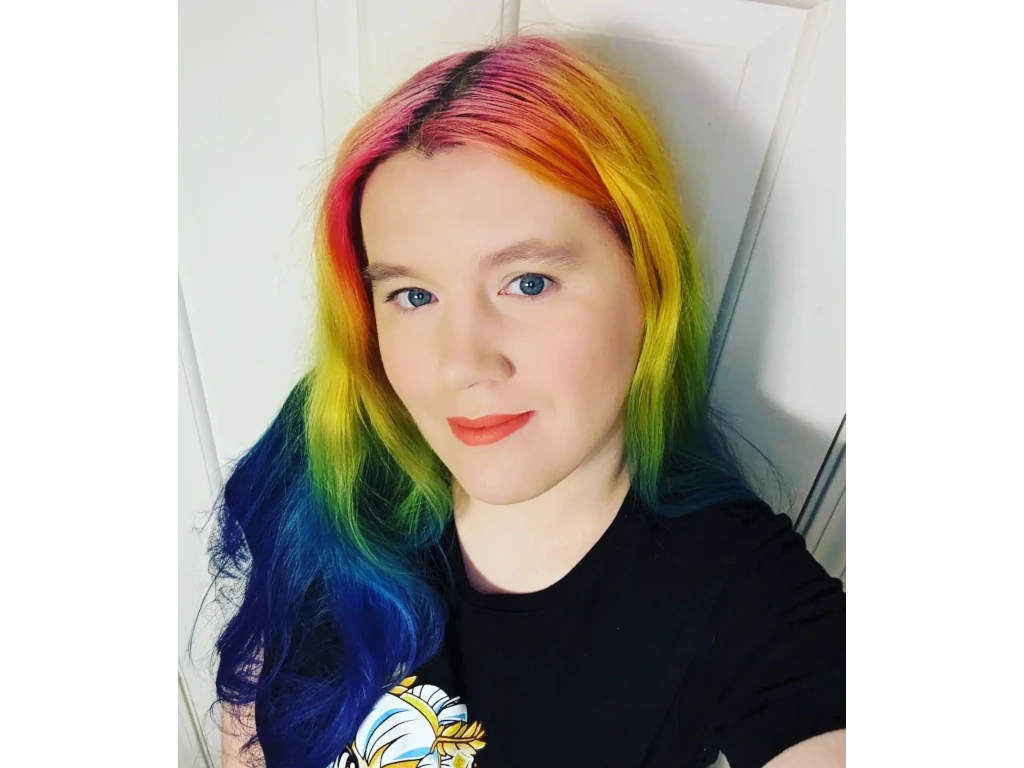 A girl with rainbow gradient hair that goes from magenta to red, to orange, to yellow, to green, to blue, and finally indigo. She has fair skin, dark red lips, bright blue eyes, and is wear a short sleeve black shirt.